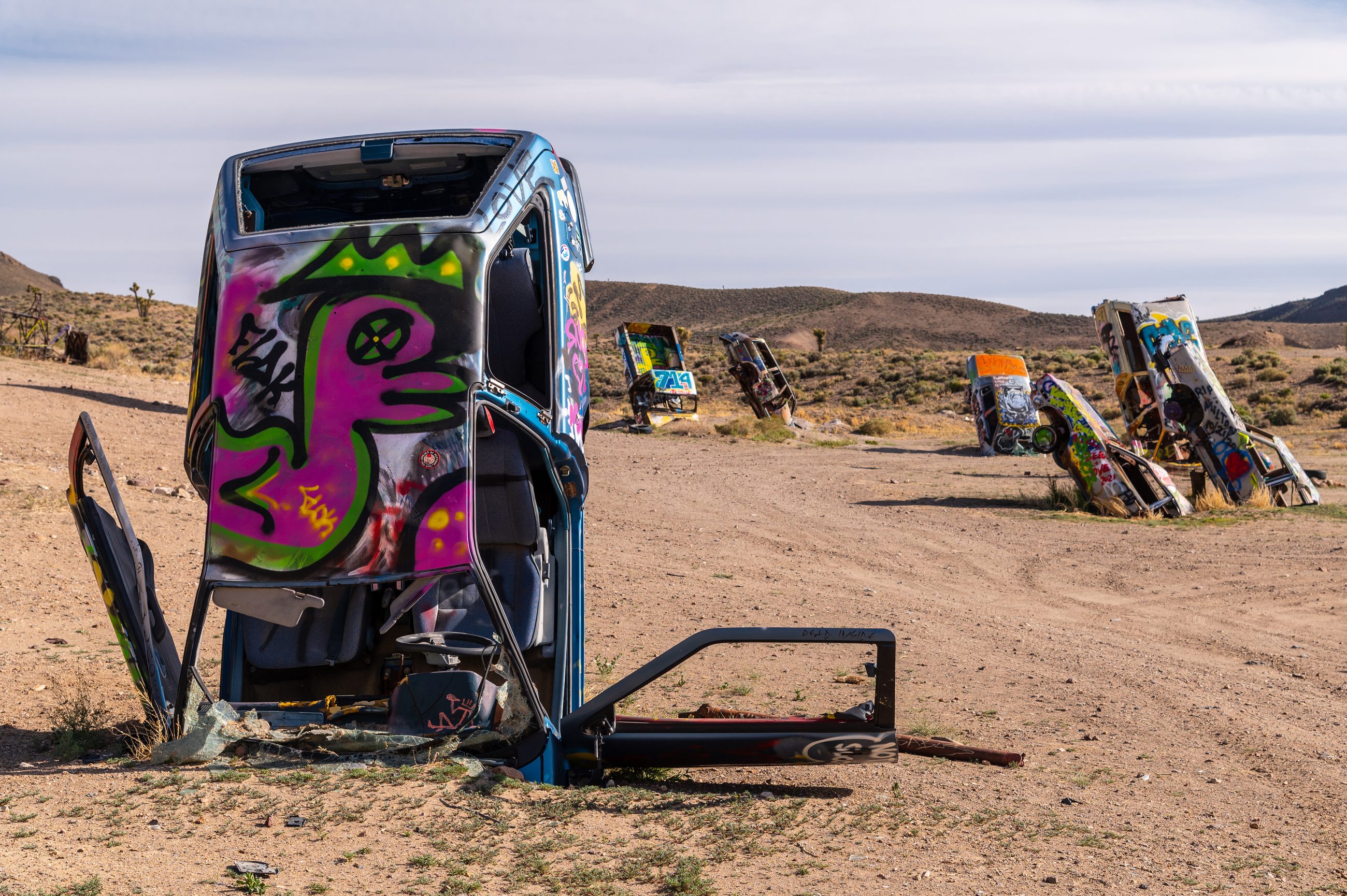 A car stood nose-down in the desert at the International Car Forest of the Last Church outside Goldfield, its exposed underside painted with a pink and green graffiti figure, its doors torn off and lying flat on the dirt beside it and its windscreen shattered across the ground. Behind it a line of upended and half-buried cars runs across the open flat, each painted in a different scheme, with Joshua trees, sagebrush and low brown hills beyond under a pale overcast sky.