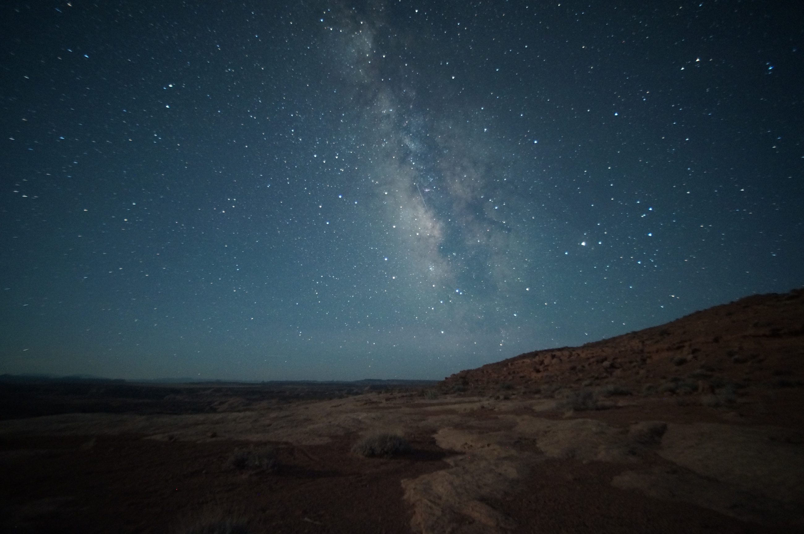 The Milky Way rising over the San Rafael Desert in Utah — bare slickrock and low scrub in the foreground, a rock slope climbing to the right, a long flat horizon and no light anywhere in the frame.