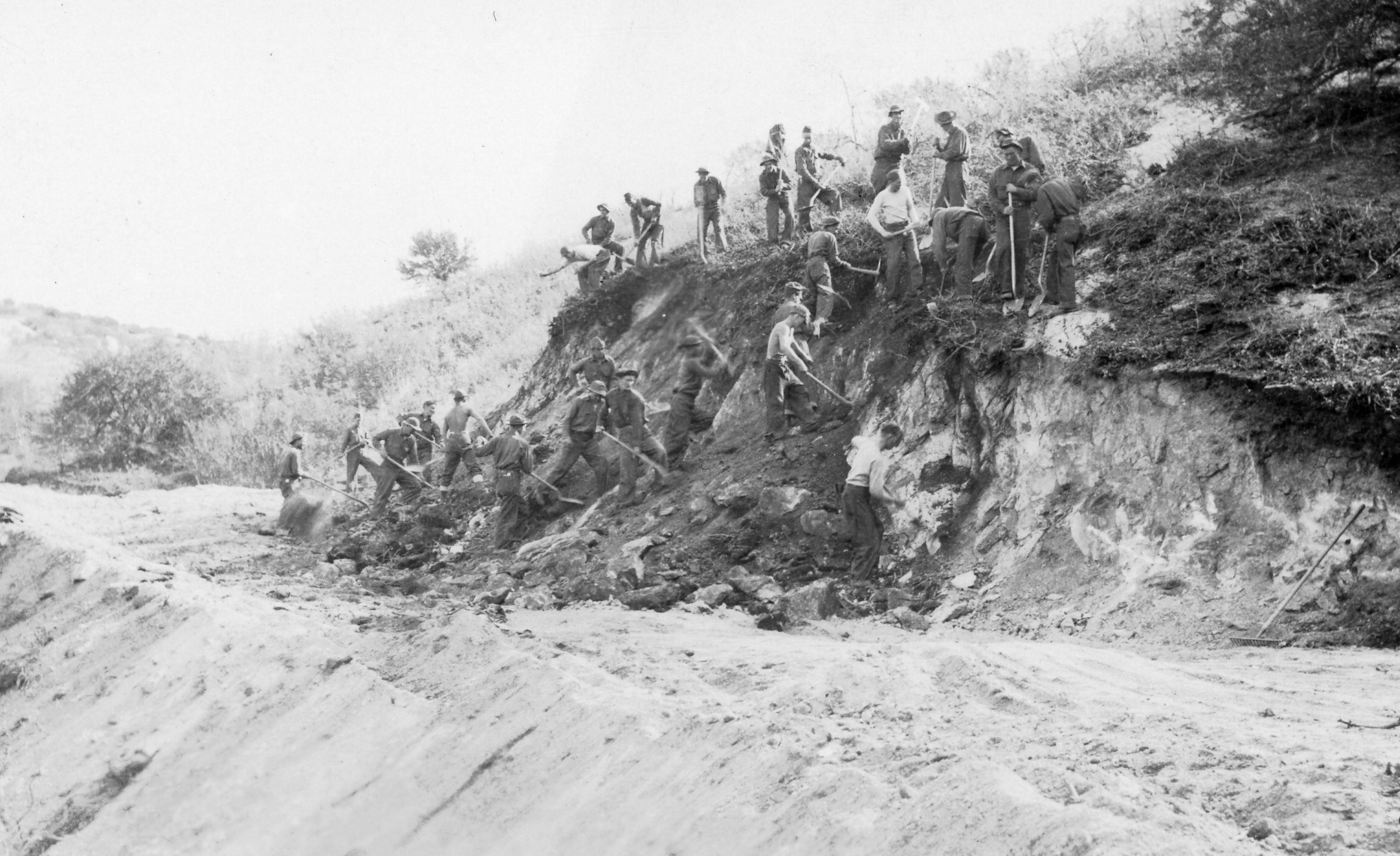 About thirty Civilian Conservation Corps enrollees working a cut bank with picks and shovels in Utah, November 1936, spread from the raw hillside face down to the freshly graded dirt roadbed below.