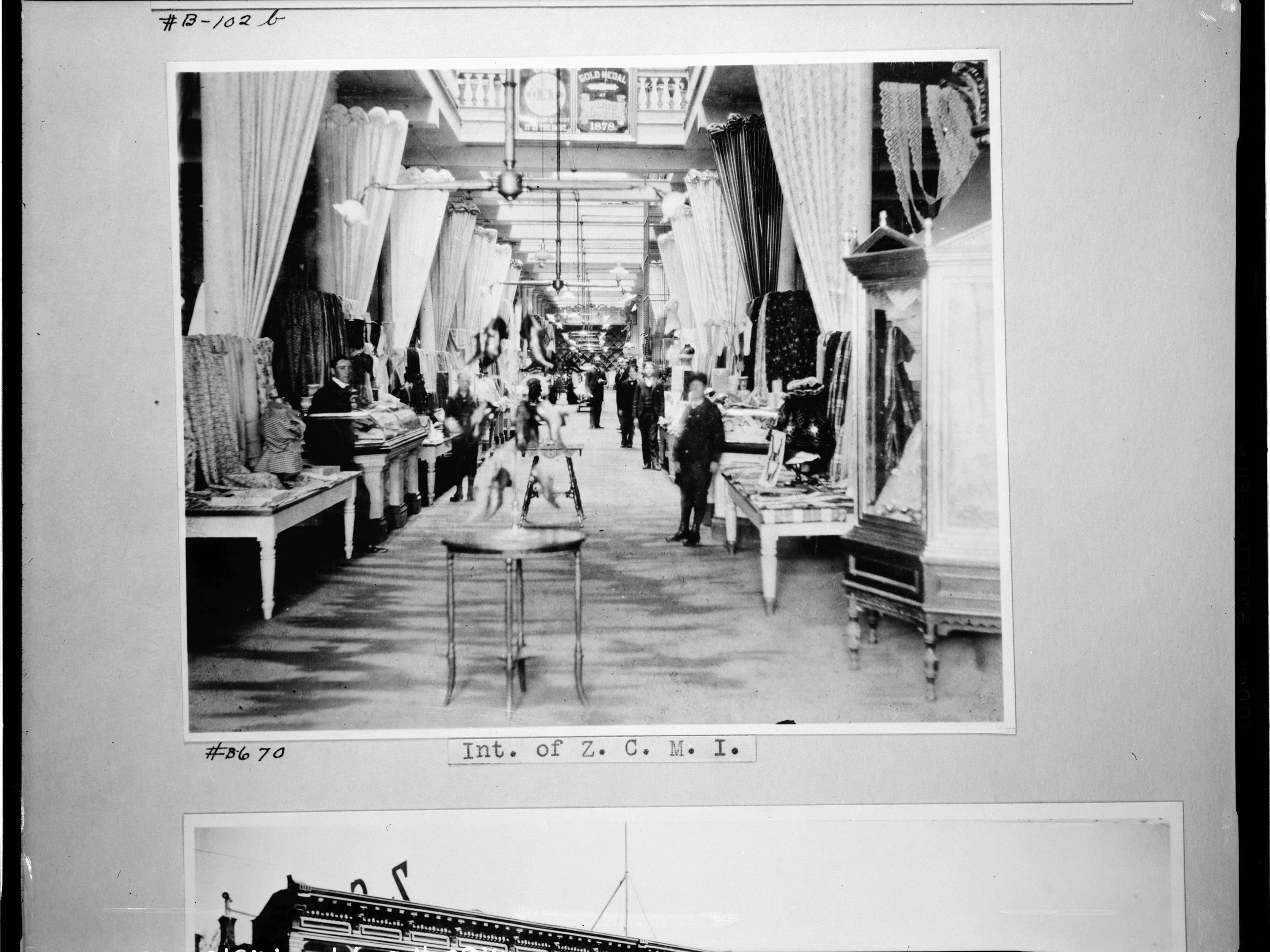 The interior of Zion's Cooperative Mercantile Institution in Salt Lake City in the late 1880s: a long aisle running between counters stacked with folded goods, iron columns and hanging fixtures down the centre, draped fabric the length of both walls, and clerks and shoppers standing among the tables