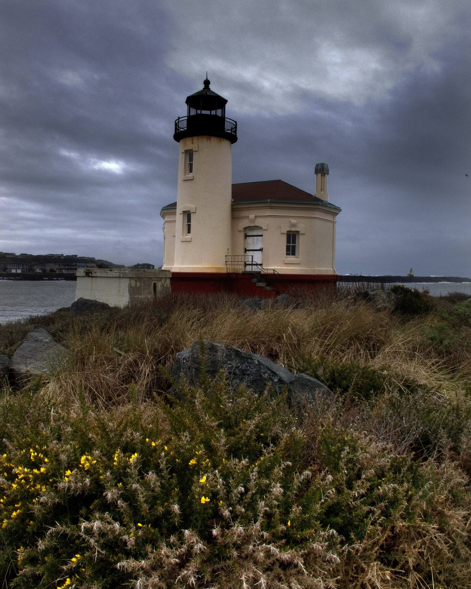 The Coquille River lighthouse at the river's mouth in Bandon, flowering scrub in the foreground under a storm sky