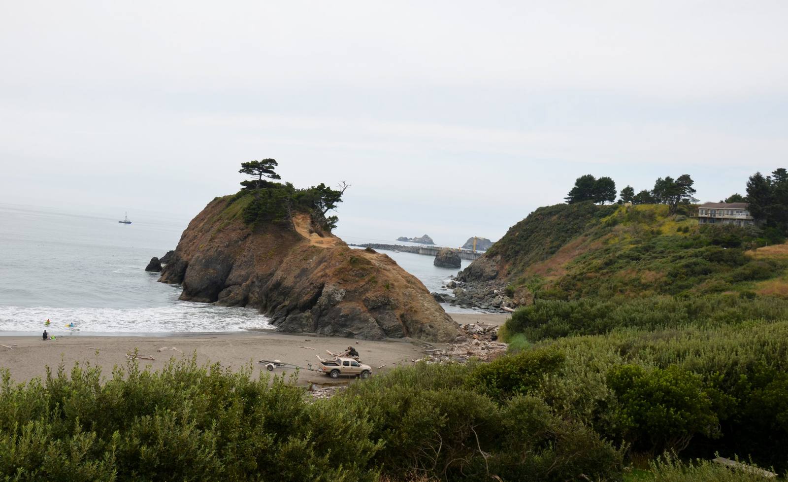 Battle Rock seen from above the beach at Port Orford: a tawny, brush-covered sea stack topped by wind-bent pines, a worn path climbing its landward side and surf breaking at its seaward foot, with a pickup and boat trailer and a lone figure on the sand below. Beyond it the harbor's dock and crane stand out on the water, and the green slope of Port Orford Heads rises at the right under an overcast sky.