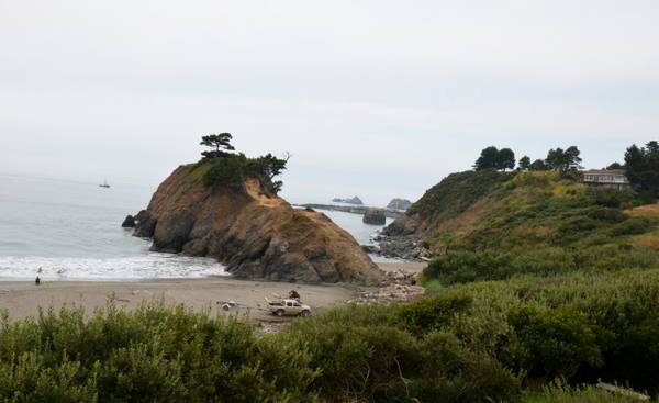 Battle Rock seen from above the beach at Port Orford: a tawny, brush-covered sea stack topped by wind-bent pines, a worn path climbing its landward side and surf breaking at its seaward foot, with a pickup and boat trailer and a lone figure on the sand below. Beyond it the harbor's dock and crane stand out on the water, and the green slope of Port Orford Heads rises at the right under an overcast sky.