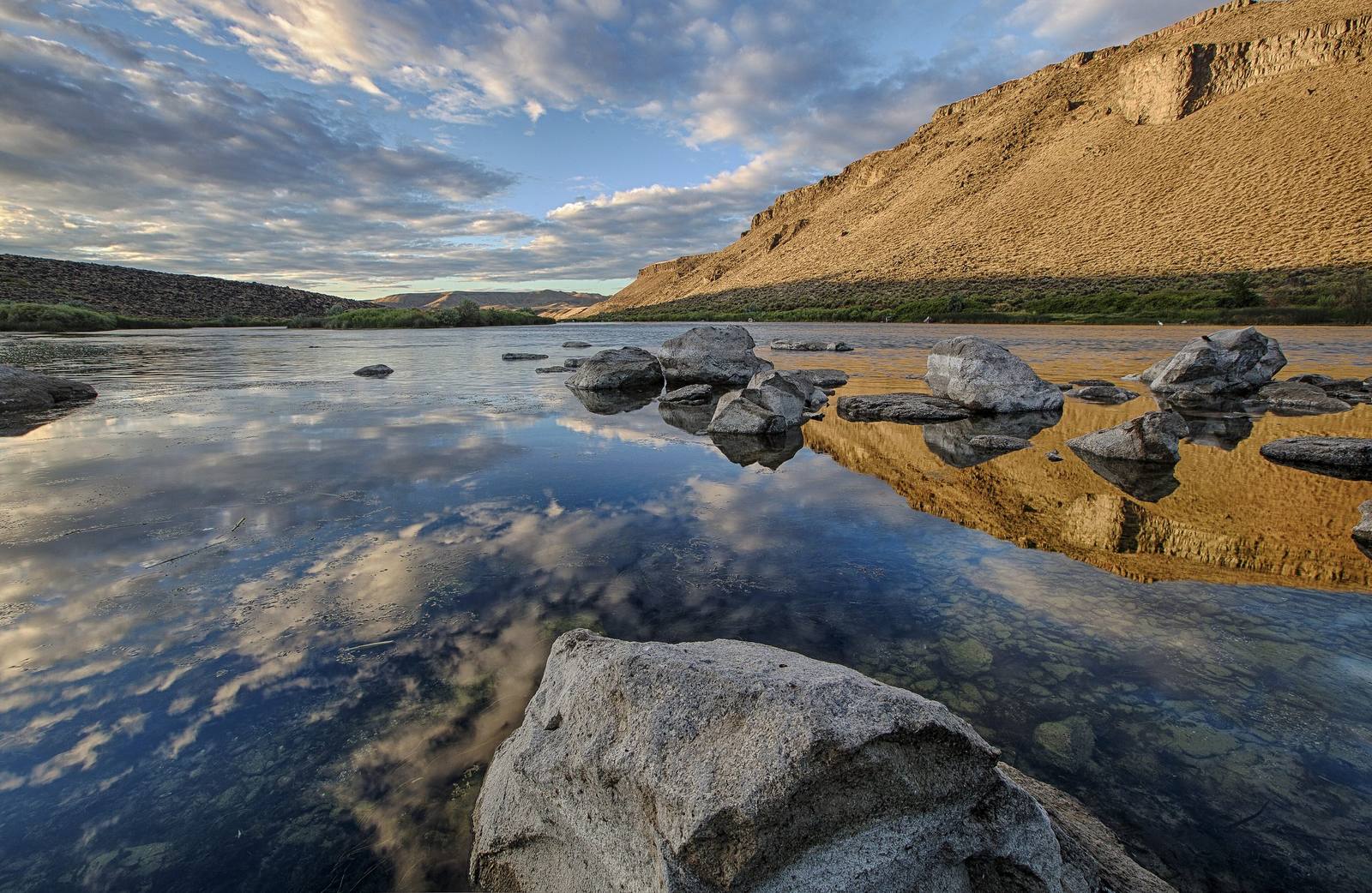 Still water of the Snake River reflecting clouds among granite boulders, beneath the canyon wall of the Birds of Prey conservation area