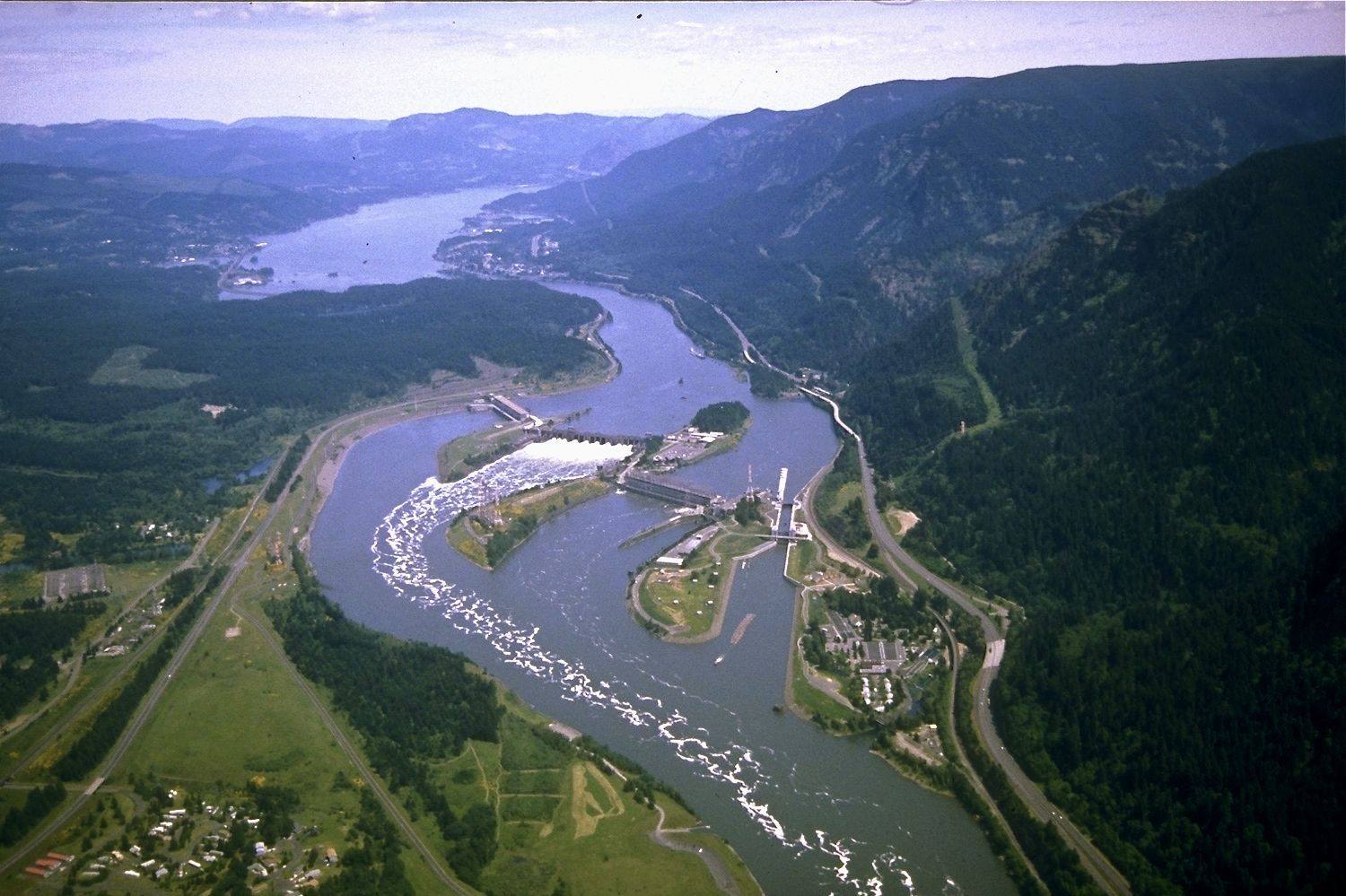 Aerial view east up the Columbia over Bonneville Lock and Dam, the spillway and powerhouses spanning the river between forested walls