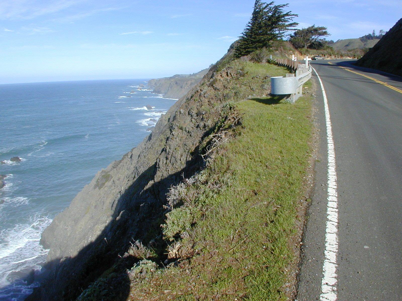 Highway 1 on the Mendocino coast running along the edge of a cliff, a guardrail on the bend ahead, the slope falling away at left to rocks and surf, with the coastline continuing south under a blue sky.