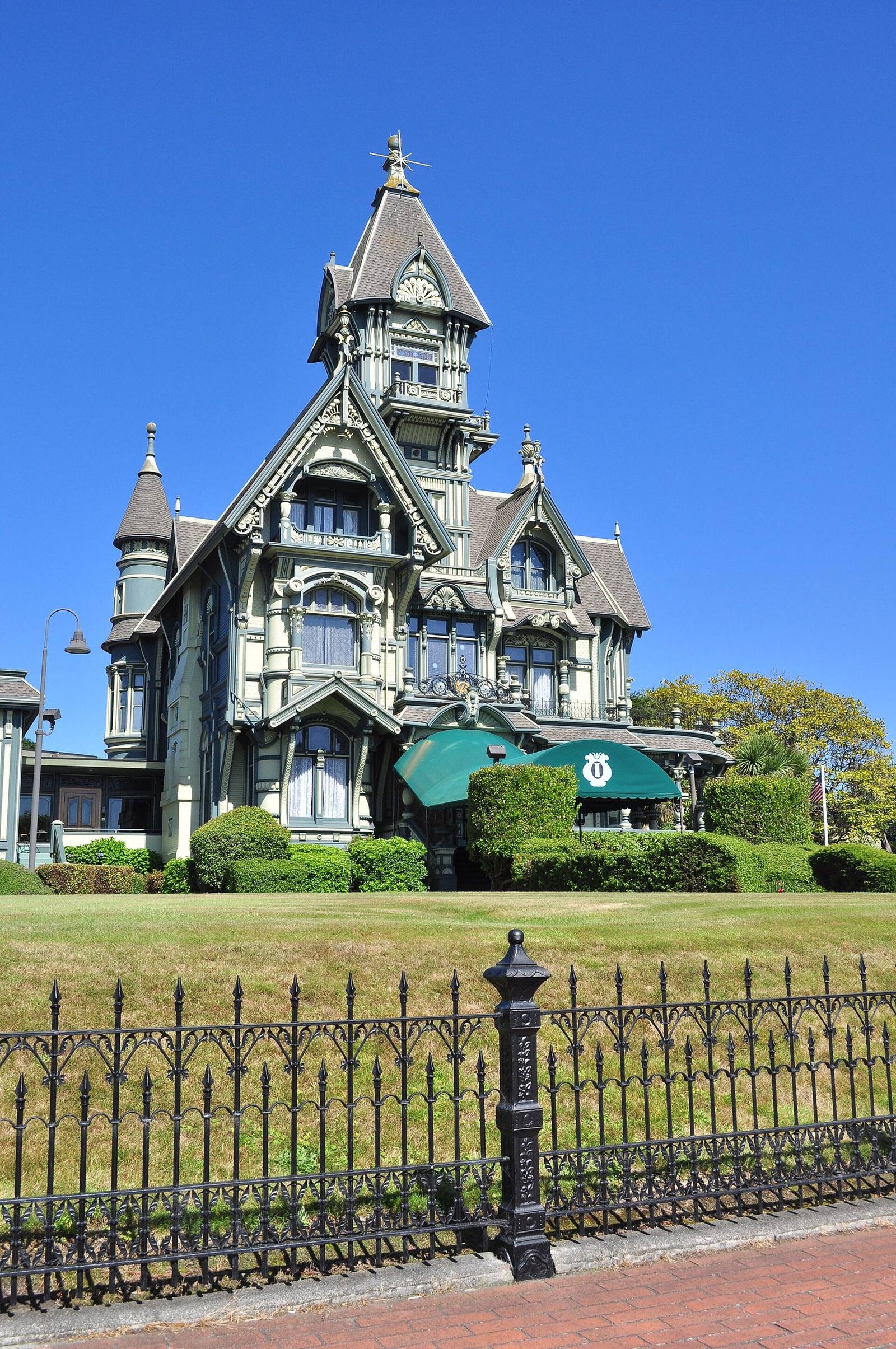 The Carson Mansion in Eureka under a clear blue sky: a green Queen Anne house of towers, gables and turned porches rising behind clipped shrubs and lawn, with an ornate iron fence along the brick sidewalk in front.