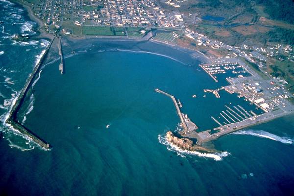 Aerial view north-northwest over Crescent City harbor: a long breakwater curling out from a rocky point, the boat basin and docks inside it at right, the curve of the bay and the town grid behind.