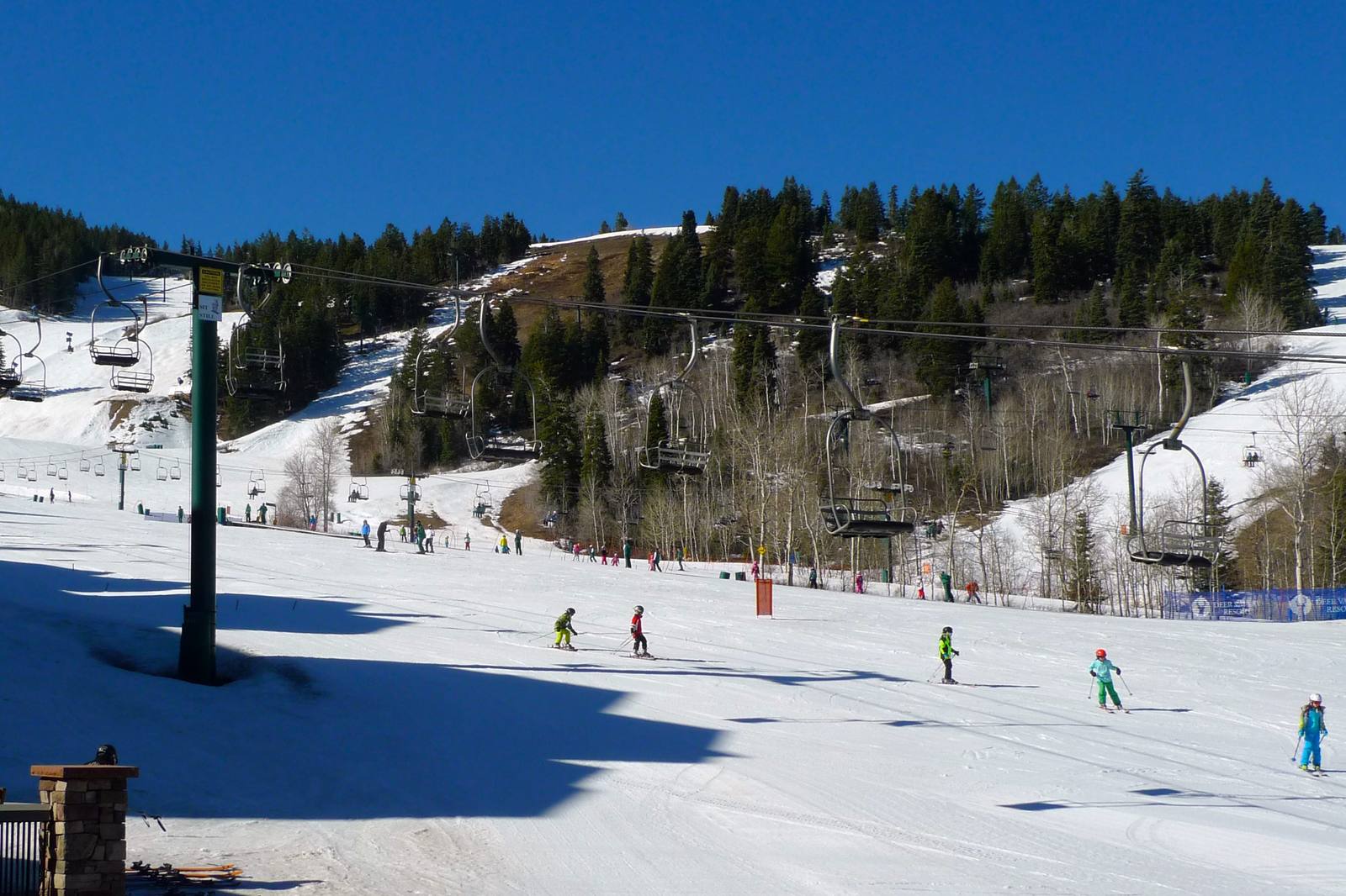 A groomed ski run in late-season sun, a broad snow apron in the foreground crossed by tree shadows, with small skiers in bright jackets and helmets strung down it in a lesson group. Three chairlift lines cross the slope behind on dark green towers, and ski runs cut pale corridors through conifer and bare aspen to a wooded ridge under cloudless blue. Bare brown ground shows through thinning snow along the run edges. A blue safety fence at right carries repeated DEER VALLEY RESORT banners.