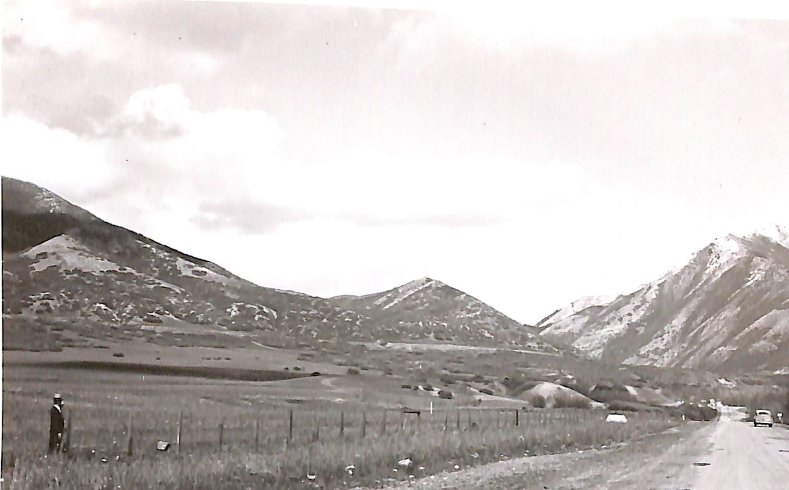 A 1946 photograph looking east up Emigration Canyon along State Road 65 — the road running away at right with a car on it, a wire fence crossing rough meadow in the foreground, a man in an overcoat and pale hat standing beside it at left, and snow-streaked ridges closing in on either side under a bright overcast sky.