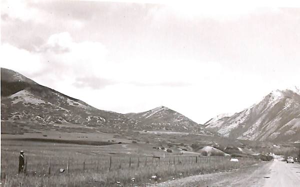 A 1946 photograph looking east up Emigration Canyon along State Road 65 — the road running away at right with a car on it, a wire fence crossing rough meadow in the foreground, a man in an overcoat and pale hat standing beside it at left, and snow-streaked ridges closing in on either side under a bright overcast sky.
