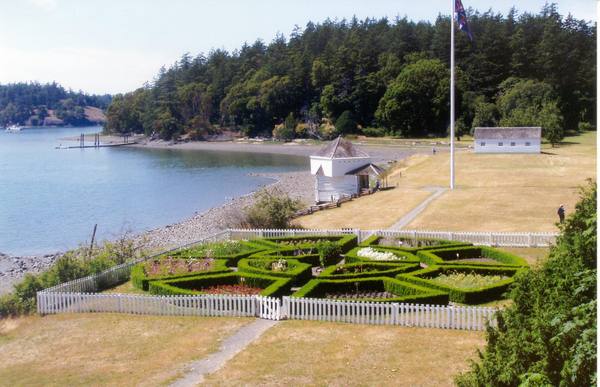 The formal garden at English Camp behind a white picket fence, with the blockhouse on the shore of Garrison Bay and the parade ground beyond