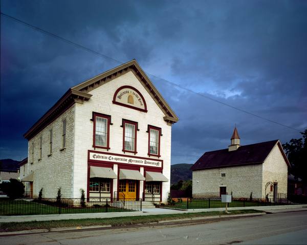 The restored Ephraim Co-op, a two-story white limestone storefront with a gold beehive and "Holiness to the Lord" painted in its pediment, under a dark storm sky beside the 1876 Relief Society granary