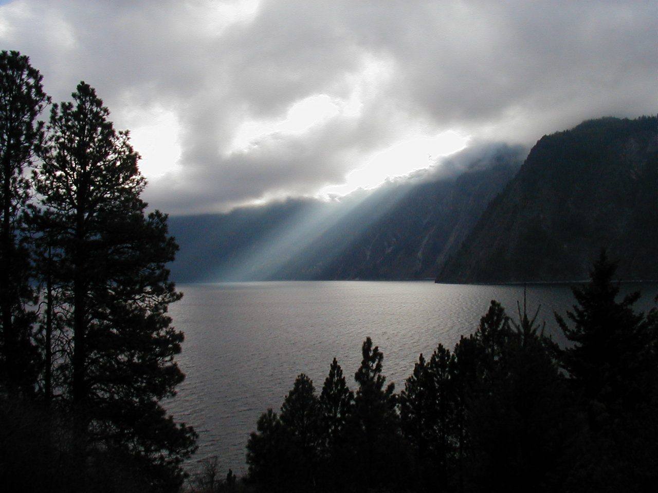 Sunlight breaking through cloud over Lake Pend Oreille below steep forested shores, seen through pines at Farragut State Park