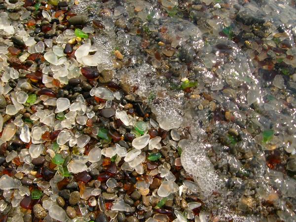 Close view of wet pebbles at Glass Beach, most of them rounded fragments of sea glass in clear, white, brown and green, with a film of surf washing over one corner.