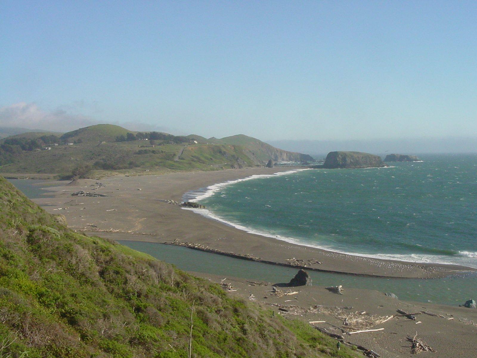 The mouth of the Russian River seen south from the Jenner cliffs: a long sandbar curving across from Jenner Beach toward Goat Rock Beach, the river backed up behind it, and the bulk of Goat Rock on the far shore with Arched Rock offshore.