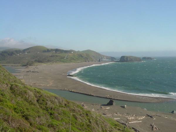 The mouth of the Russian River seen south from the Jenner cliffs: a long sandbar curving across from Jenner Beach toward Goat Rock Beach, the river backed up behind it, and the bulk of Goat Rock on the far shore with Arched Rock offshore.