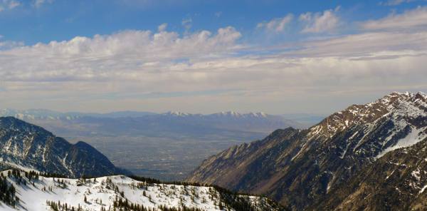 Snow-covered ridges and evergreens falling away from a summit above Little Cottonwood Canyon, with the hazy, snowless Salt Lake Valley and its street grid spread far below.