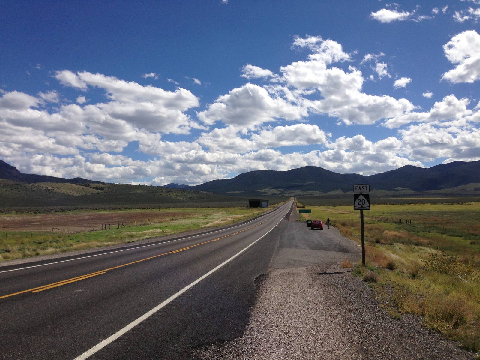 Utah State Route 20 running east as a two-lane highway toward low dark mountains on the horizon, an EAST 20 route shield standing on the gravel shoulder, rangeland either side under scattered cumulus