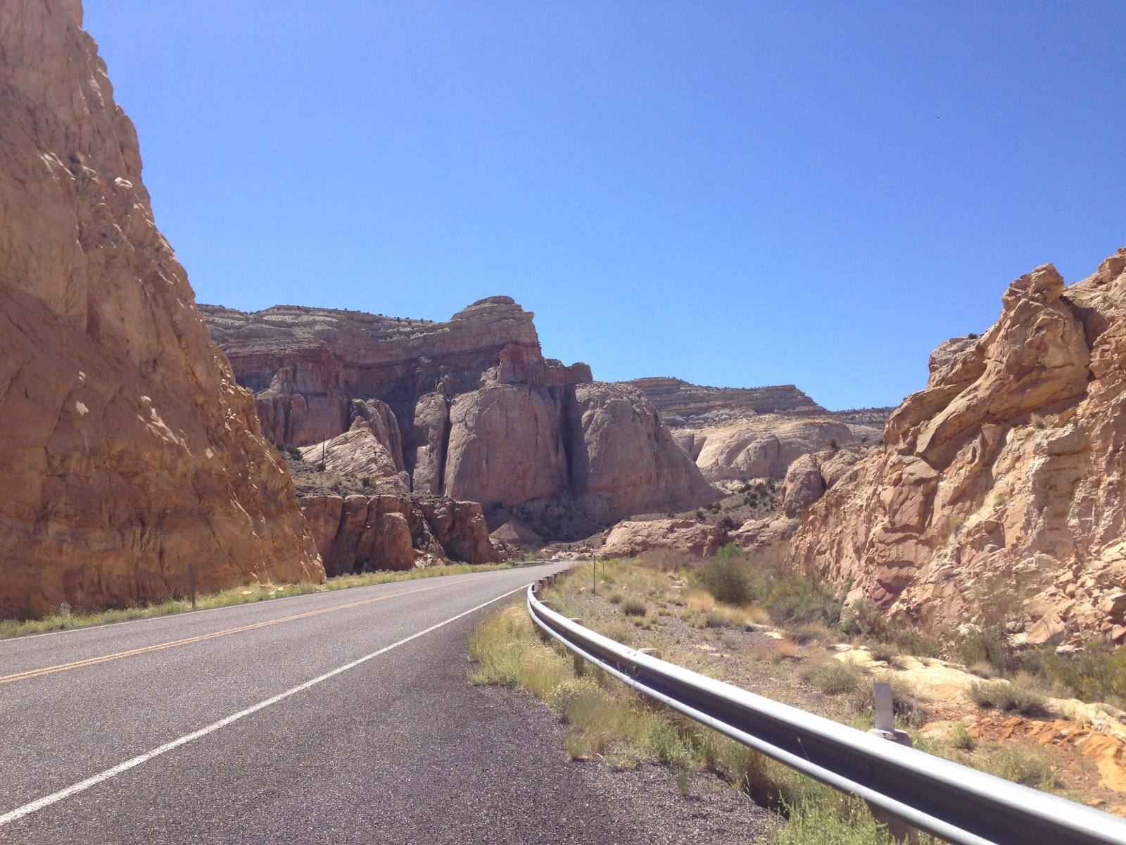 Highway 24 curving right between sandstone walls, a double yellow line down the pavement and a steel guardrail running along the right shoulder above dry brush. Banded cliffs and pale rounded domes of gold and white rock stand ahead and to either side under a cloudless deep blue sky. The Fremont River, which the road follows through this gorge, is not visible in the frame.