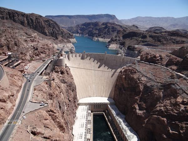 Hoover Dam curving between the cliffs of Black Canyon, Lake Mead behind it, the powerhouse wings and the Colorado River in the gorge below, and the highway switchbacking down to the crest.