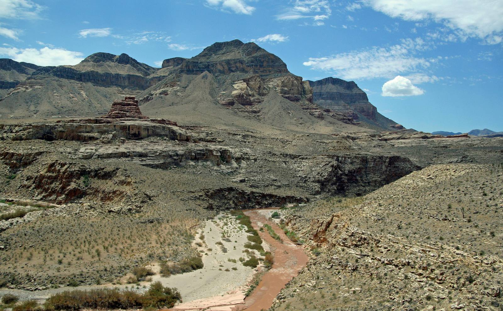 The Virgin River running muddy red along the floor of its gorge in the far northwest corner of Arizona, threading a broad pale wash of sand and gravel scattered with green brush. Tilted grey and buff limestone beds rise in stepped ledges on both sides, a small red-capped butte stands left of centre, and a high banded ridge closes the view under blue sky and scattered cumulus. Interstate 15 is the vantage point rather than anything in the picture: the photograph was taken from the highway itself.