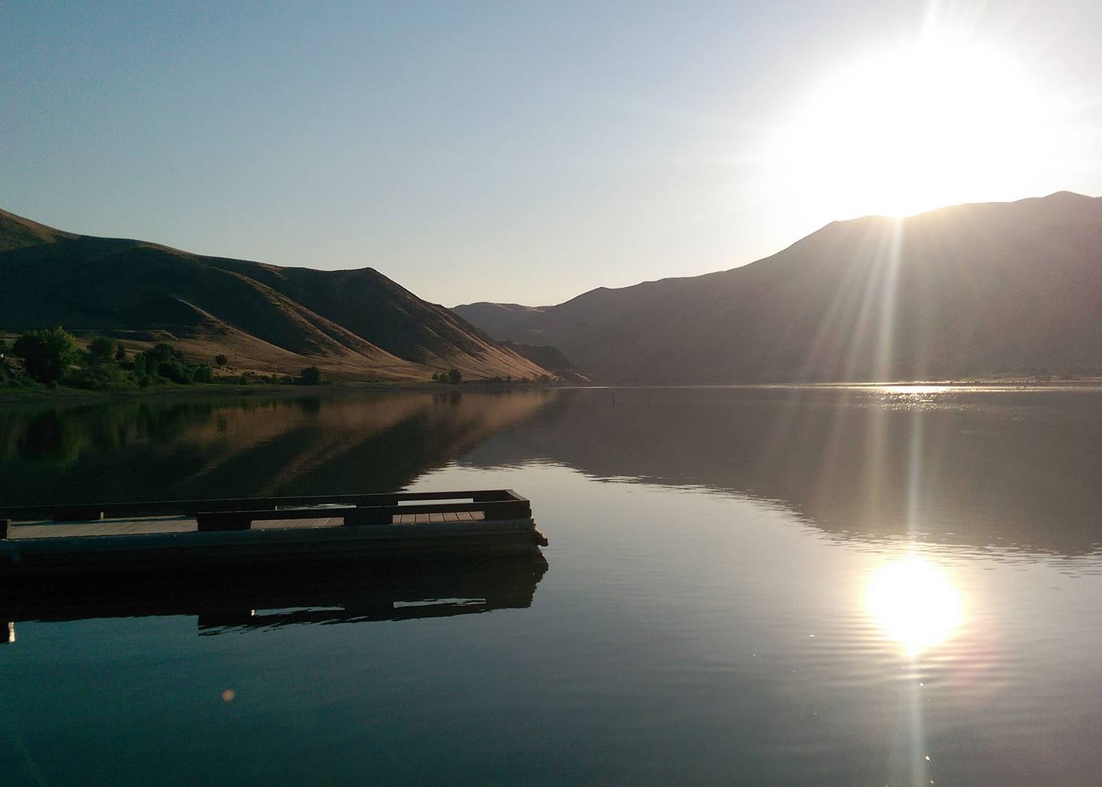 The Snake River at Farewell Bend, now the slack water of Brownlee Reservoir, early on a summer morning: the low sun flares over brown grassy hills at the right and lays a bright path across the calm water, a wooden dock floats in the near water at the left, and trees stand along the far shore beneath the hills.