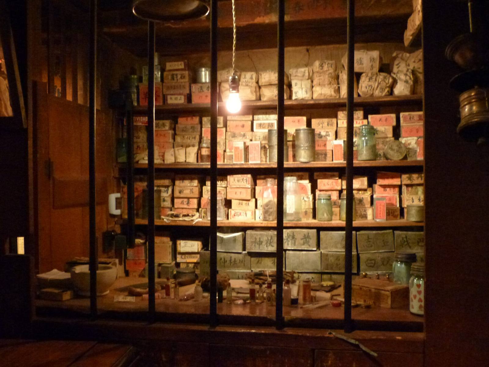 Shelves of packaged herbs and remedies behind the apothecary window at Kam Wah Chung, lit by a single bulb