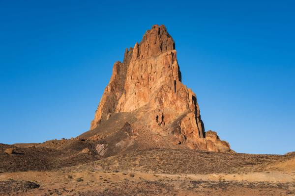 Agathla Peak — Aghaałą́ — the eroded volcanic neck standing seven miles north of Kayenta beside US-163, rising in a single jagged mass of dark breccia more than fifteen hundred feet above the desert, its spires and gullies picked out in low sun and its flanks falling into a long scree apron over pale banded ground. Bare rock and sparse scrub run to the foreground under a cloudless sky. The peak is sacred to the Diné and is not climbed. The town of Kayenta is not in the frame.