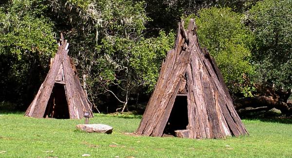 Two conical houses of leaning redwood bark slabs standing on a mown clearing at Kule Loklo, a flat stone in the grass between them and oak woodland behind.