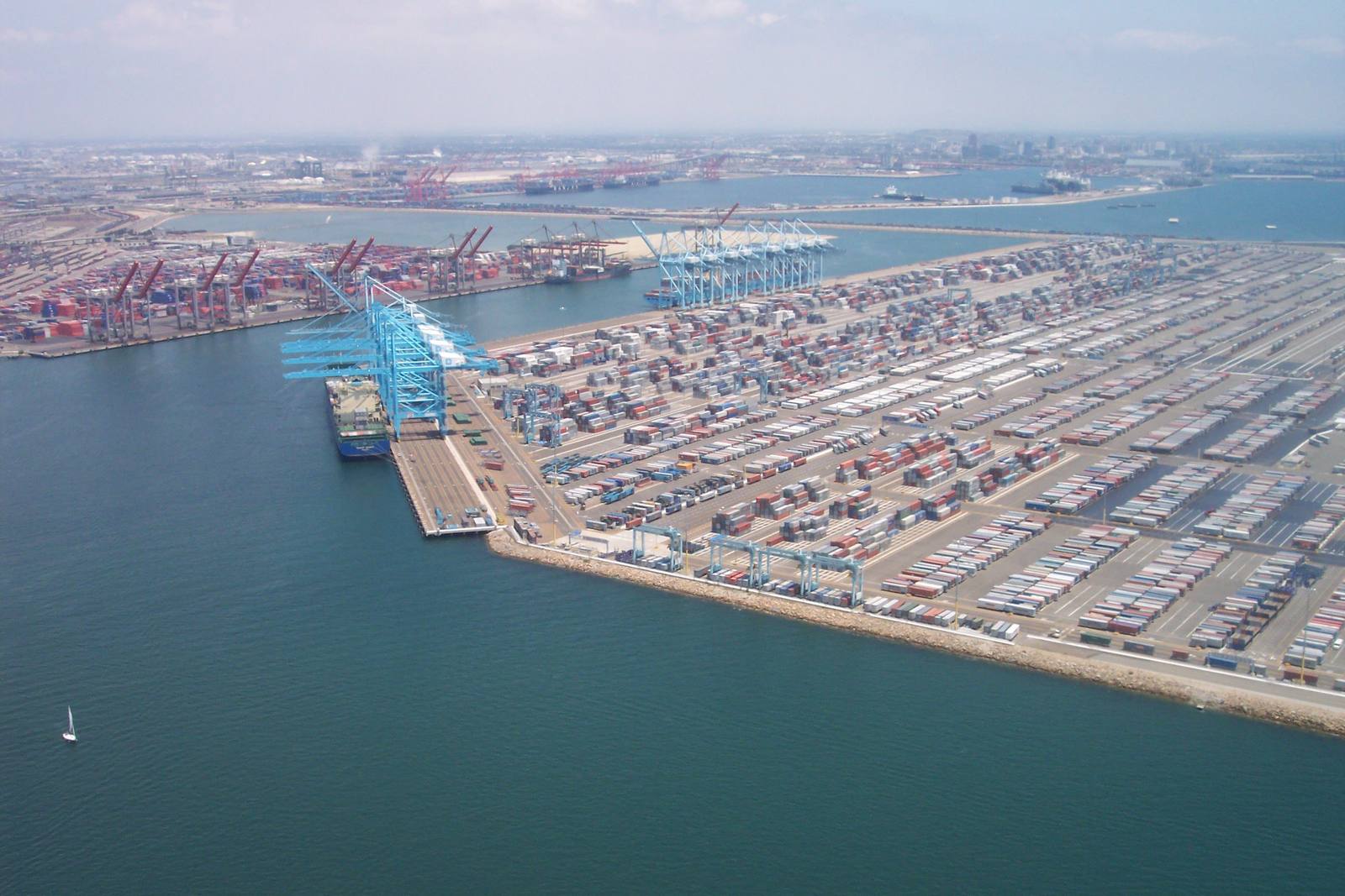 Aerial view of San Pedro Bay: container cranes and rows of stacked cargo standing on made ground, with the breakwater and the open Pacific beyond.