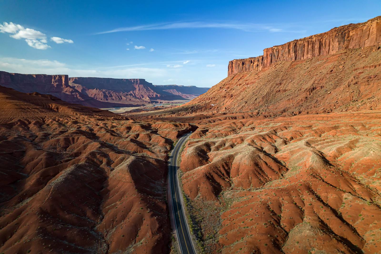 The northern end of the La Sal Loop Road seen from the air, where it comes down out of the hills into Castle Valley from Highway 128 — two lanes with a double yellow line curving up the middle of the frame through deeply rilled red slopes, one vehicle on the pavement at the top of the rise. A tall mesa capped in dark red rock fills the right side above long fluted talus, and beyond the road the ground opens into a wide valley with a green river bottom and a line of flat-topped mesas running back to the horizon under thin cloud. Arches, Canyonlands and the town of Moab — the region's headline places — are all out of frame.