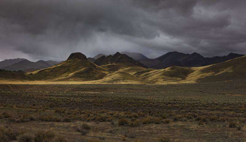 Basin and Range country under a breaking storm — heavy dark overcast filling the top of the frame with rain trailing beneath it, and a low shaft of sun raking gold across a line of grassy foothills below. Darker ranges stand behind them in shadow, layering back to a hazy ridge at the left. In the foreground a broad sagebrush flat lies in shade, a wire fence on leaning posts running across it from edge to edge.