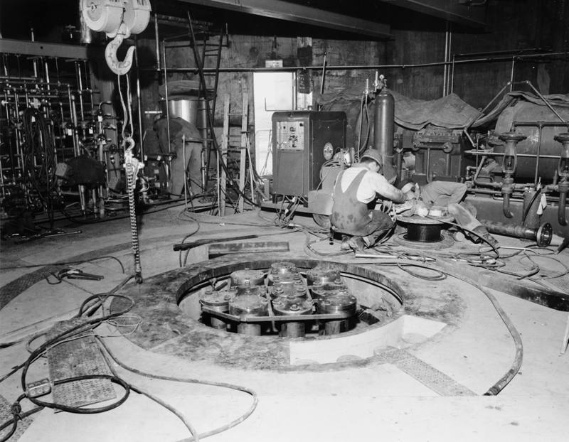 The head of the SL-1 reactor in place in the floor of the reactor building in March 1958, nearly three years before the accident, its control-rod drive housings in the open pit and workmen with welding gear beyond