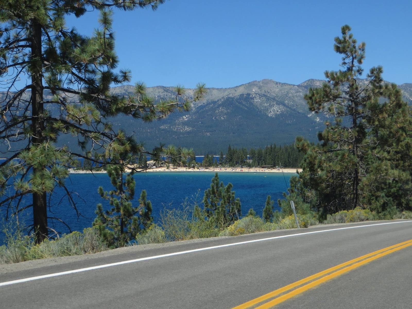 State Route 28's pavement crosses the foreground, its double yellow centre line and white edge line sharp in the sun. Beyond a bank of sagebrush and pines, Lake Tahoe runs deep blue over a pale turquoise shallow shelf to a long crescent of sand where small figures, umbrellas and low buildings sit among the trees. The forested Carson Range rises behind to a bare granite crest.