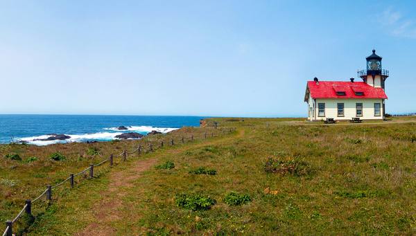 Point Cabrillo Light Station on its grassy headland: the white building with a red roof and the lantern rising from its seaward end, a fenced path running toward it and the ocean and offshore rocks at left.
