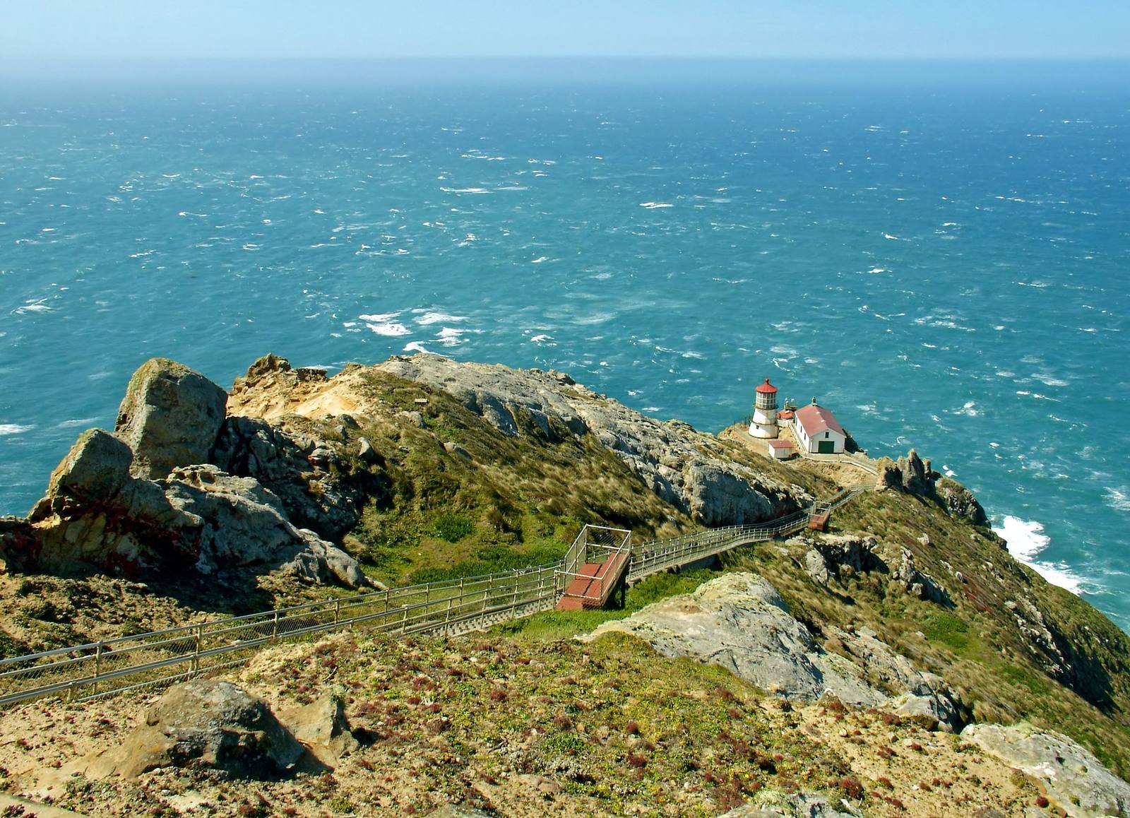 Point Reyes Lighthouse seen from the headland above: the small white tower and red-roofed building sit on a shelf low on the rocky point, a long stairway zigzagging down the slope toward them, with the open Pacific beyond.