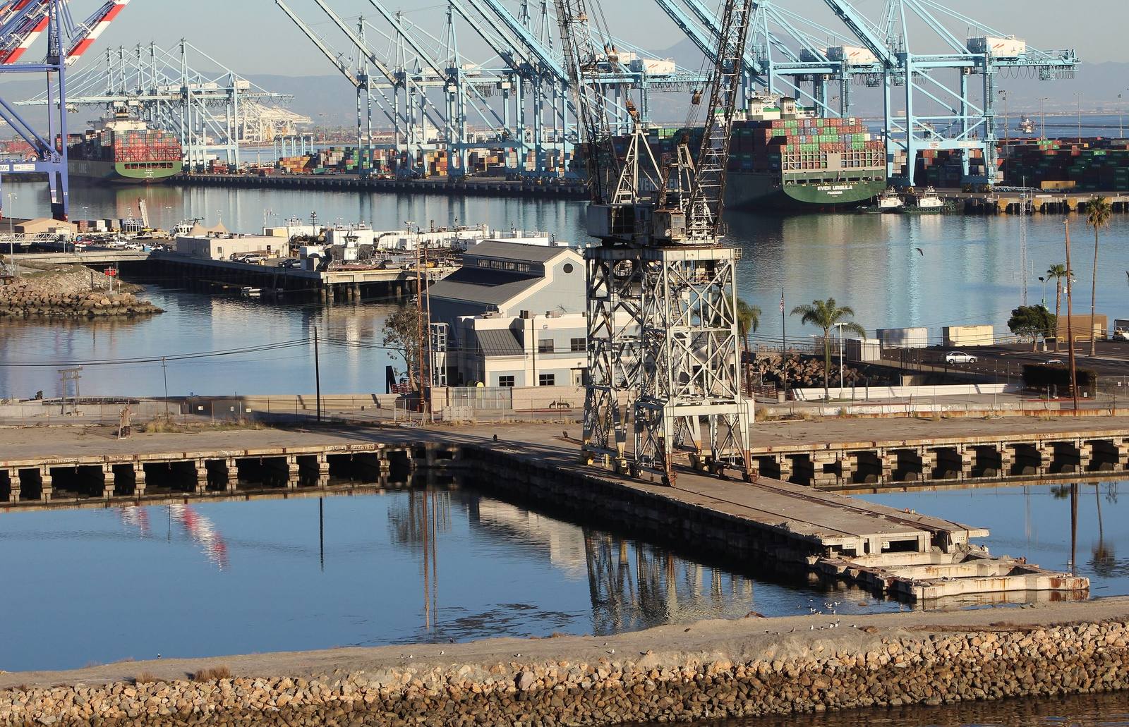 Old timber docks and a derrick in the foreground of the Port of Los Angeles, with container cranes and a berthed ship behind.