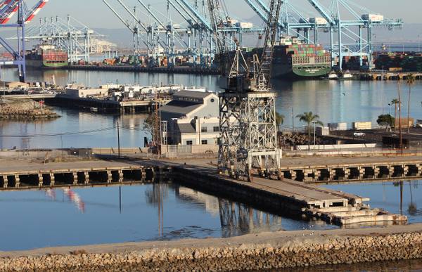 Old timber docks and a derrick in the foreground of the Port of Los Angeles, with container cranes and a berthed ship behind.