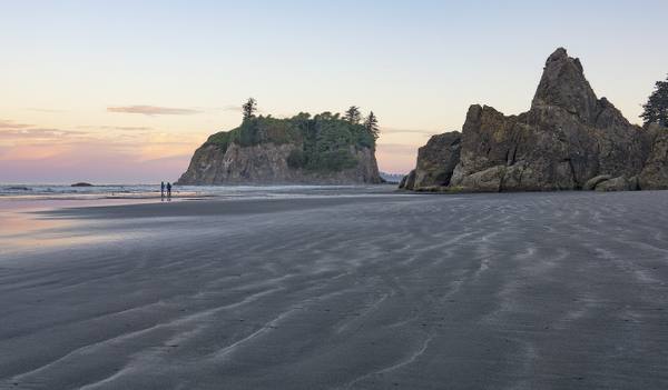 Ruby Beach at low tide, rippled wet sand running out to tree-topped Abbey Island and a jagged sea stack, two people walking at the water's edge