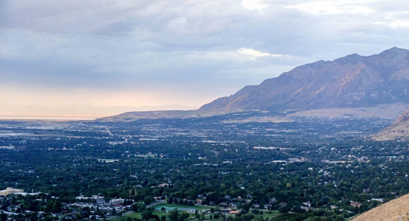 Ogden spread under tree canopy across the valley floor at dusk, the bare wall of the Wasatch rising on the right and the Great Salt Lake pale on the horizon to the left.