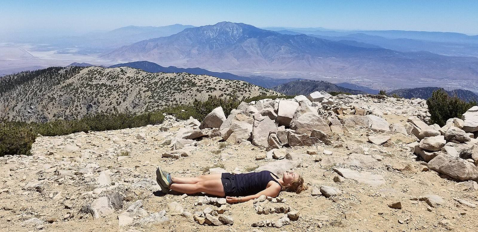 The bare rock summit of San Gorgonio Mountain above the tree line, a hiker resting among the stones, San Jacinto Peak across the pass beyond.