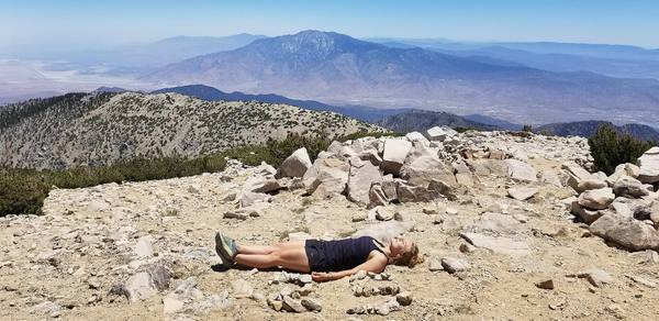 The bare rock summit of San Gorgonio Mountain above the tree line, a hiker resting among the stones, San Jacinto Peak across the pass beyond.