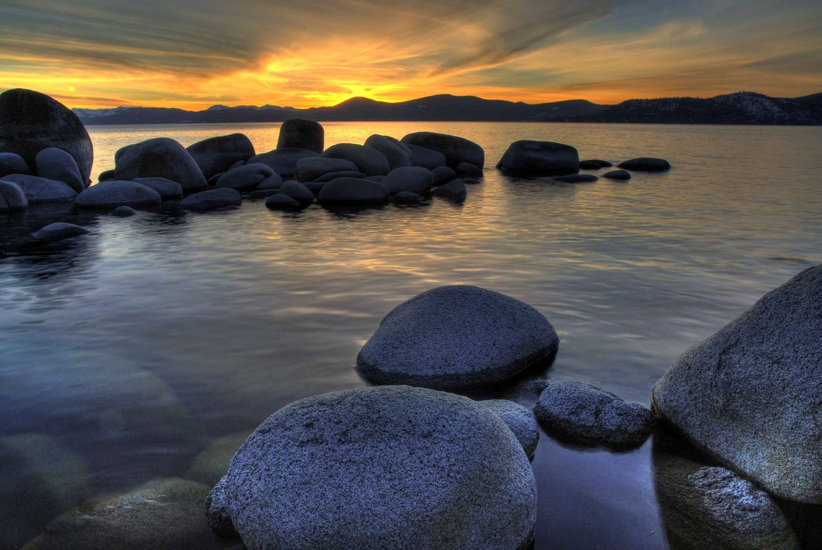 Granite boulders standing in glassy still water at Sand Harbor at sunset, an orange and gold sky above the dark, snow-flecked ridgeline across Lake Tahoe.
