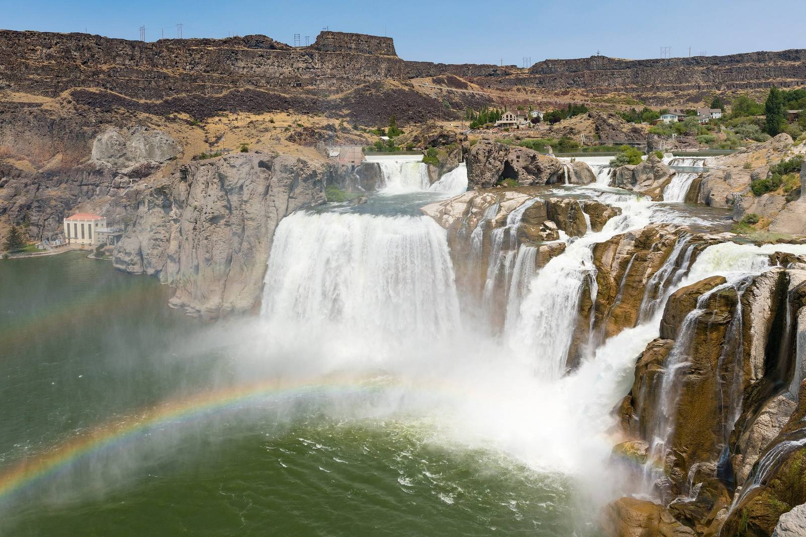 Shoshone Falls pouring over its basalt rim in several channels, a rainbow in the spray and the power plant at left