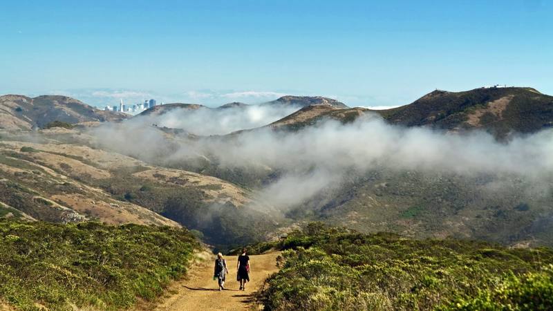 Two hikers on the Coastal Trail along Coyote Ridge in the Marin Headlands, bare grassy hills folding away in front of them, fog filling the valleys, and the towers of San Francisco showing through a gap on the horizon.