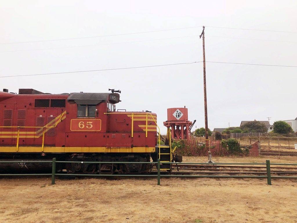 A red and yellow California Western diesel locomotive numbered 65 standing on the tracks at the Fort Bragg depot, a water tank and low buildings beyond it under a white sky.