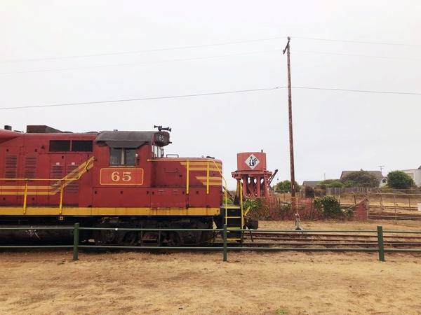 A red and yellow California Western diesel locomotive numbered 65 standing on the tracks at the Fort Bragg depot, a water tank and low buildings beyond it under a white sky.