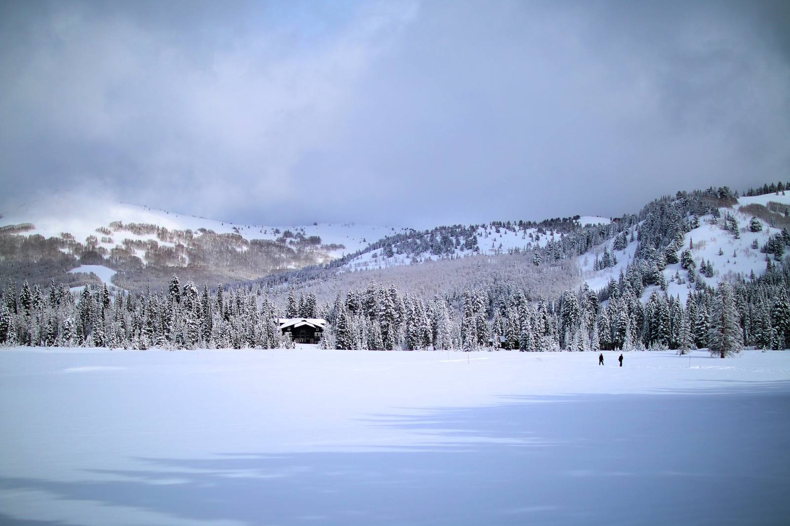 The Solitude Nordic Center at Silver Lake under deep snow — a dark timber lodge standing at the treeline beyond an unbroken snowfield, with snow-laden conifers running back up the slope, cut ski runs showing on the mountainside above, and two small figures crossing the open snow. Low cloud sits along the ridge.
