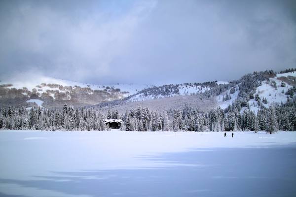 The Solitude Nordic Center at Silver Lake under deep snow — a dark timber lodge standing at the treeline beyond an unbroken snowfield, with snow-laden conifers running back up the slope, cut ski runs showing on the mountainside above, and two small figures crossing the open snow. Low cloud sits along the ridge.